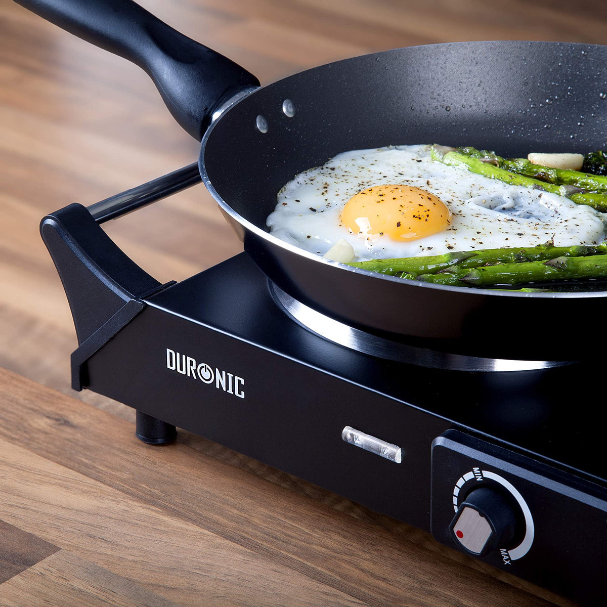 Duronic HP1 BK Hornillo Eléctrico de Camping 1500 W | Mostrando un primer plano del hornillo eléctrico en una superficie de madera, con una sartén encima donde se cocinan un huevo frito y espárragos, destacando el panel frontal con la marca, la luz piloto y el control giratorio de temperatura