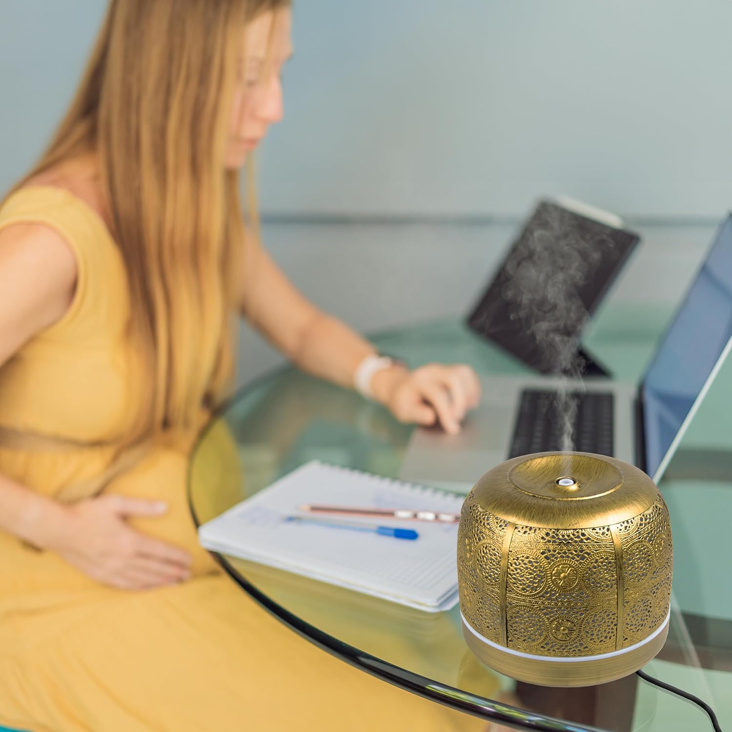 Duronic AD43 Difusor de Aromas y Brumizador de Aceites Esenciales Mostrando el difusor de aromaterapia dorado de 500 ml en funcionamiento, emitiendo bruma sobre una mesa de vidrio junto a una persona trabajando con un ordenador portátil y notas