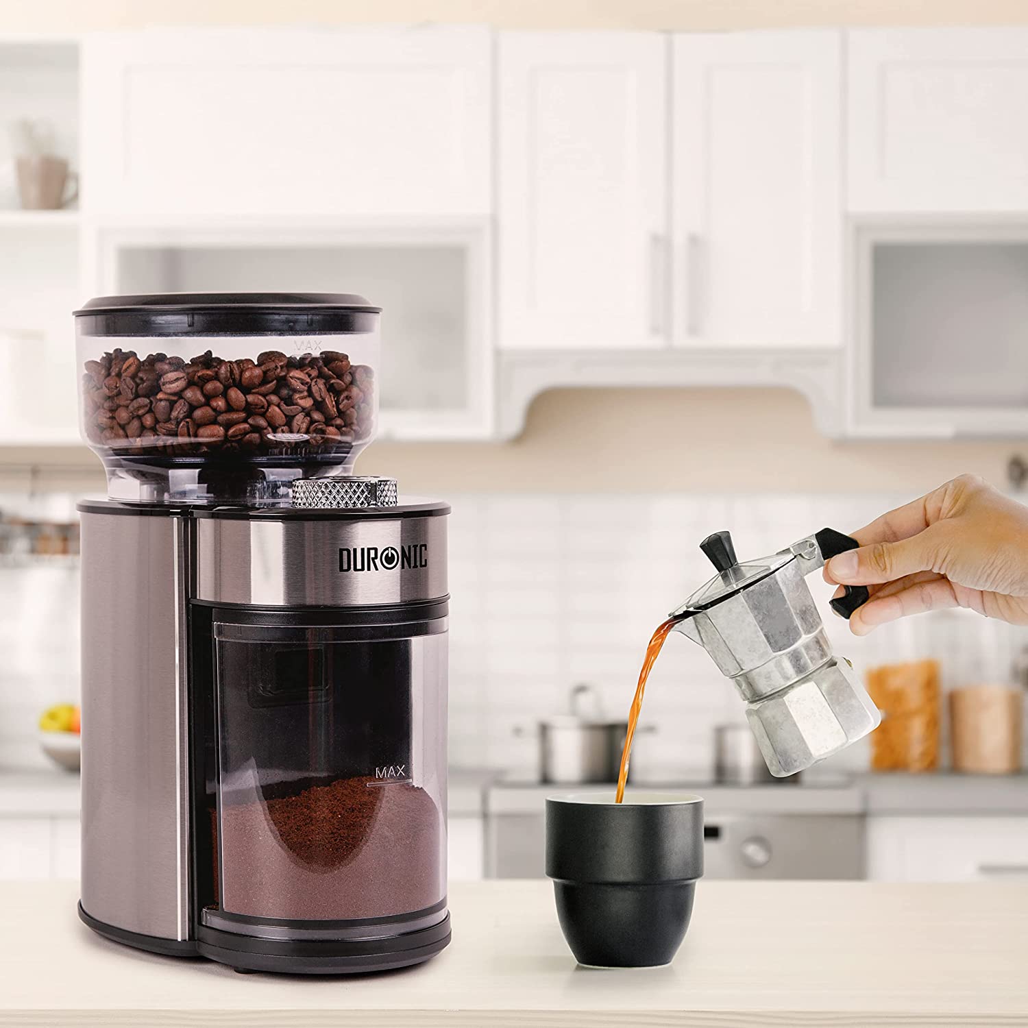 Duronic BG200 Molinillo de café eléctrico | Mostrando el molinillo de café eléctrico sobre una encimera en una cocina, con granos en el depósito superior y café molido en el inferior, mientras una mano vierte café desde una cafetera italiana en una taza de color negro