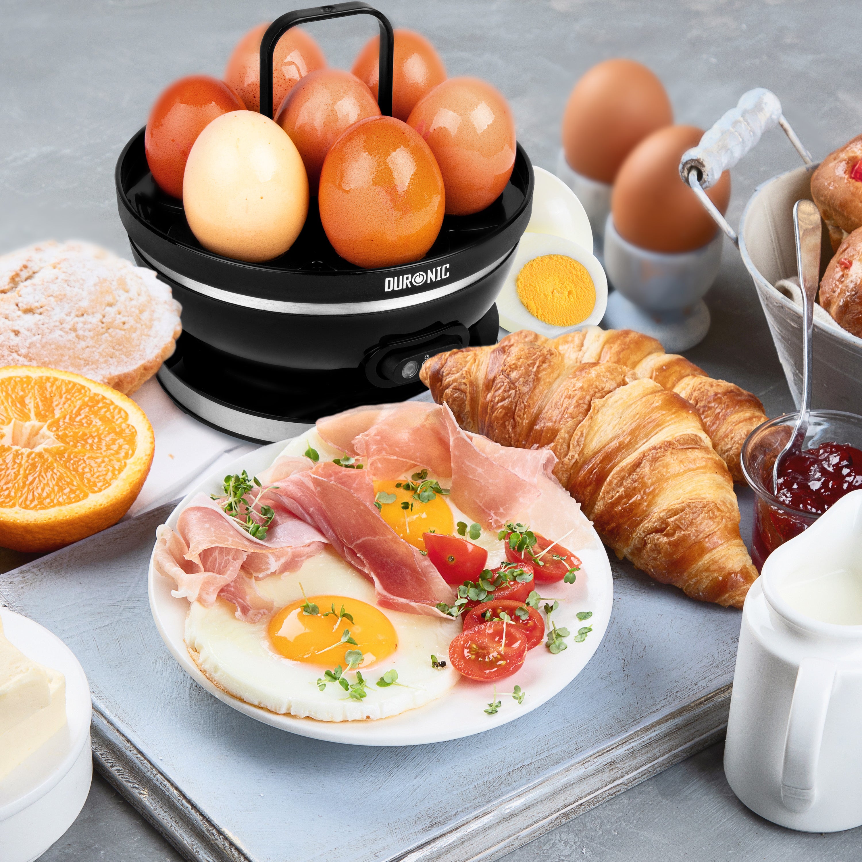 Duronic EB27 BK Hervidor de Huevos Eléctrico 400 W para 7 Huevos | Mostrando el hervidor de huevos eléctrico negro sobre una mesa con un desayuno completo que incluye huevos cocidos en su bandeja soporte, un plato con huevos fritos y jamón, croissants, fruta y mermelada