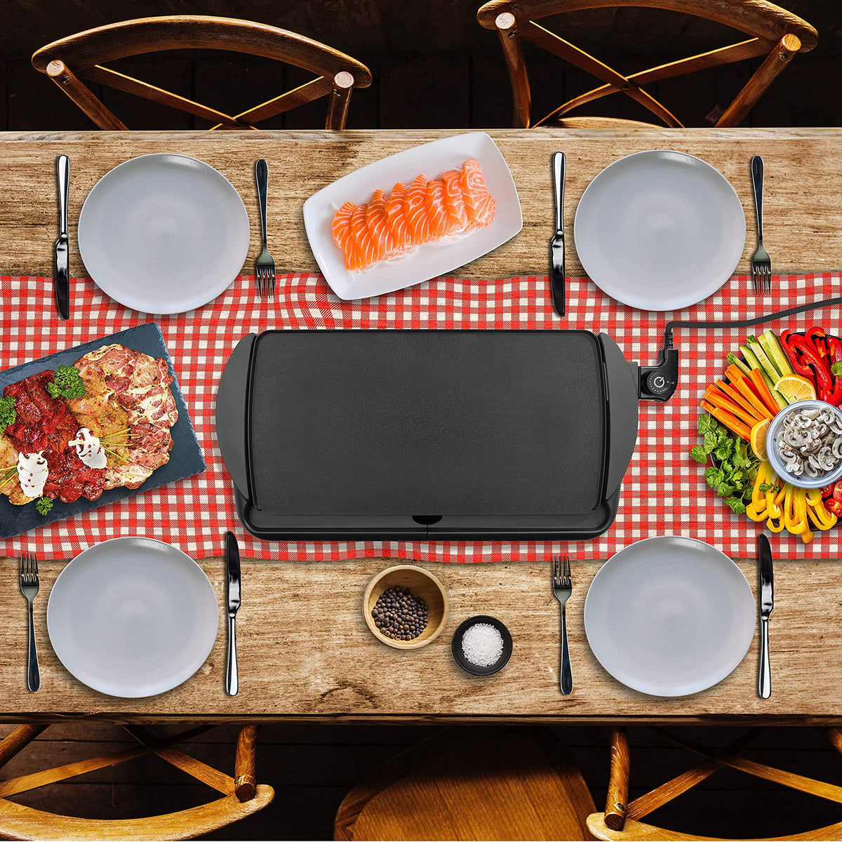 Duronic GP20 Plancha Eléctrica Grill Mostrando la plancha colocada en el centro de una mesa de comedor decorada con mantel de cuadros, rodeada de platos y diversos ingredientes frescos como salmón, carne y verduras listos para cocinar