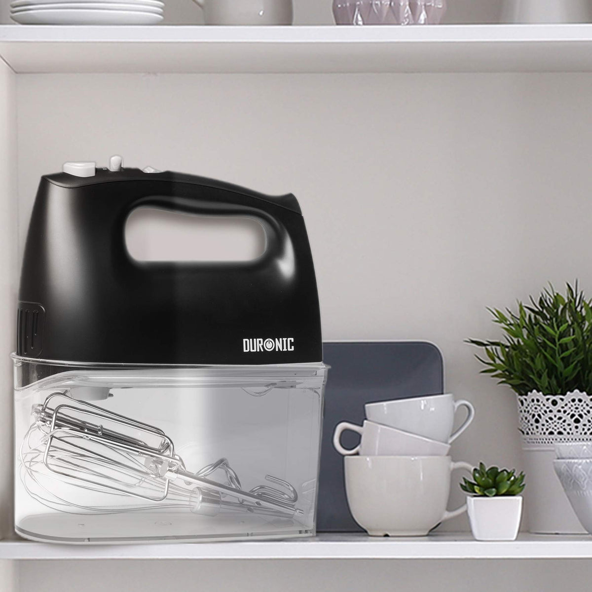 Duronic HM4 - Batidora de Varillas y Amasadora | Mostrando la batidora en color negro almacenada de forma compacta sobre su base transparente, con los accesorios guardados en su interior y colocada en un estante de cocina junto a tazas y una planta pequeña