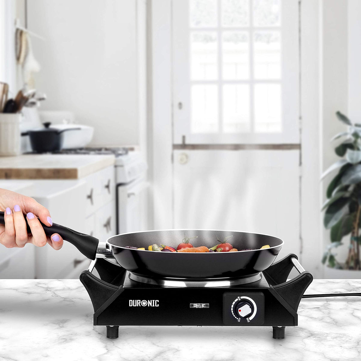 Duronic HP1 BK Hornillo Eléctrico de Camping 1500 W | Mostrando el hornillo eléctrico colocado sobre una encimera de cocina de mármol blanco, mientras una mano sujeta el mango de una sartén con verduras salteadas que desprende vapor