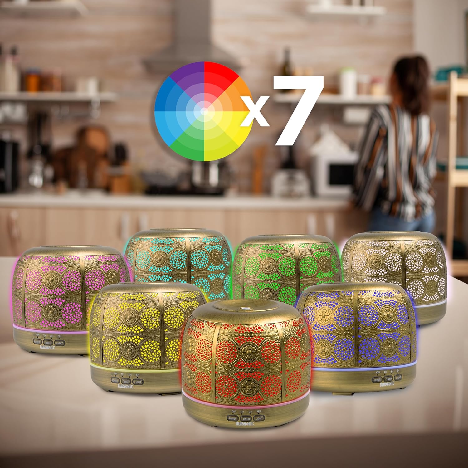 Duronic AD43 Difusor de Aromas y Brumizador de Aceites Esenciales Mostrando múltiples unidades del difusor de aromaterapia dorado con carcasa metálica calada dispuestas sobre una encimera de cocina, cada una iluminada con un color LED diferente para destacar sus 7 opciones de iluminación ambiental