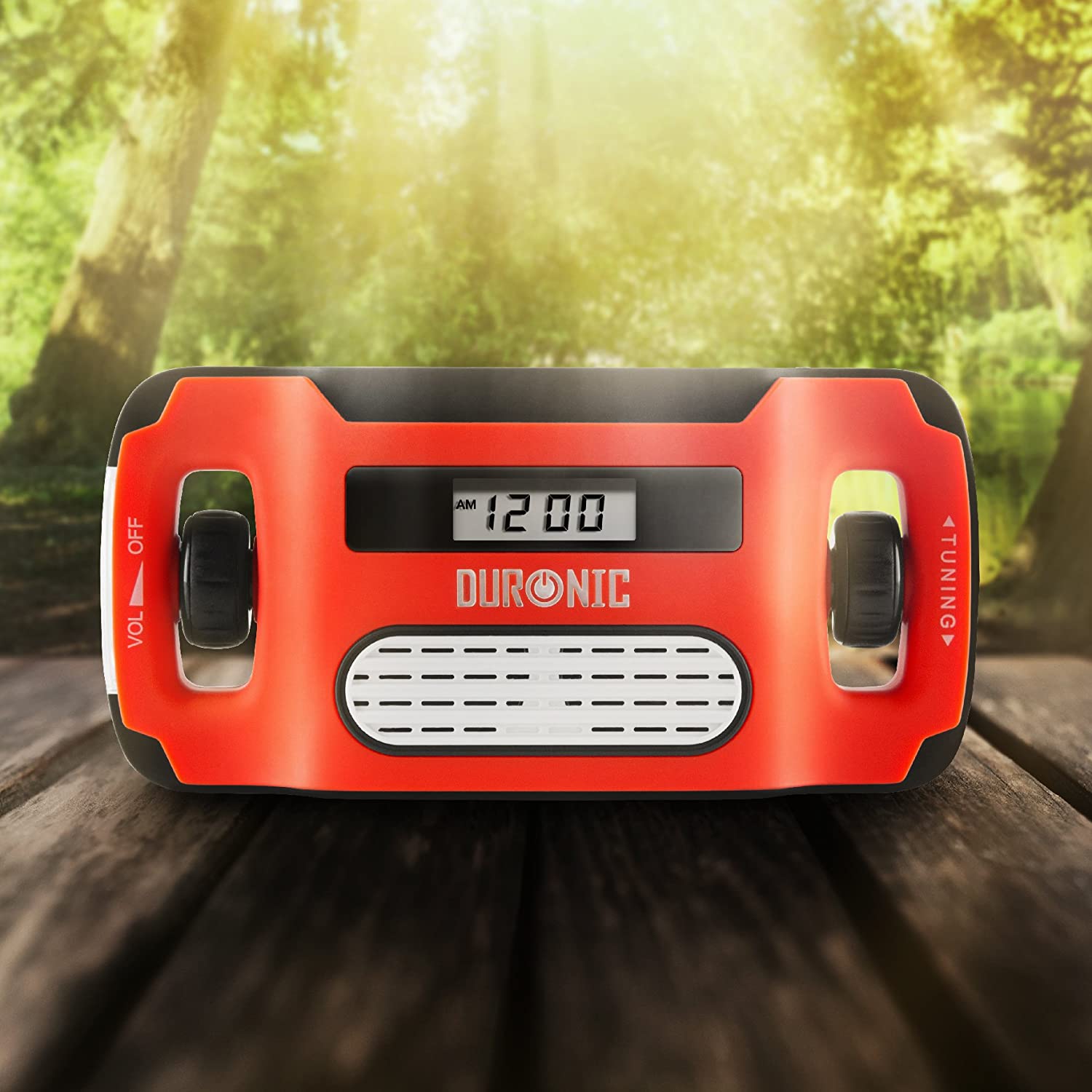 Duronic Apex Radio Solar de Emergencia con Manivela y Batería Recargable | Mostrando la radio portátil de emergencia sobre una superficie de madera al aire libre con un fondo natural de árboles iluminados por el sol, destacando su diseño en color rojo y negro, la pantalla digital con reloj y la marca Duronic en el panel frontal