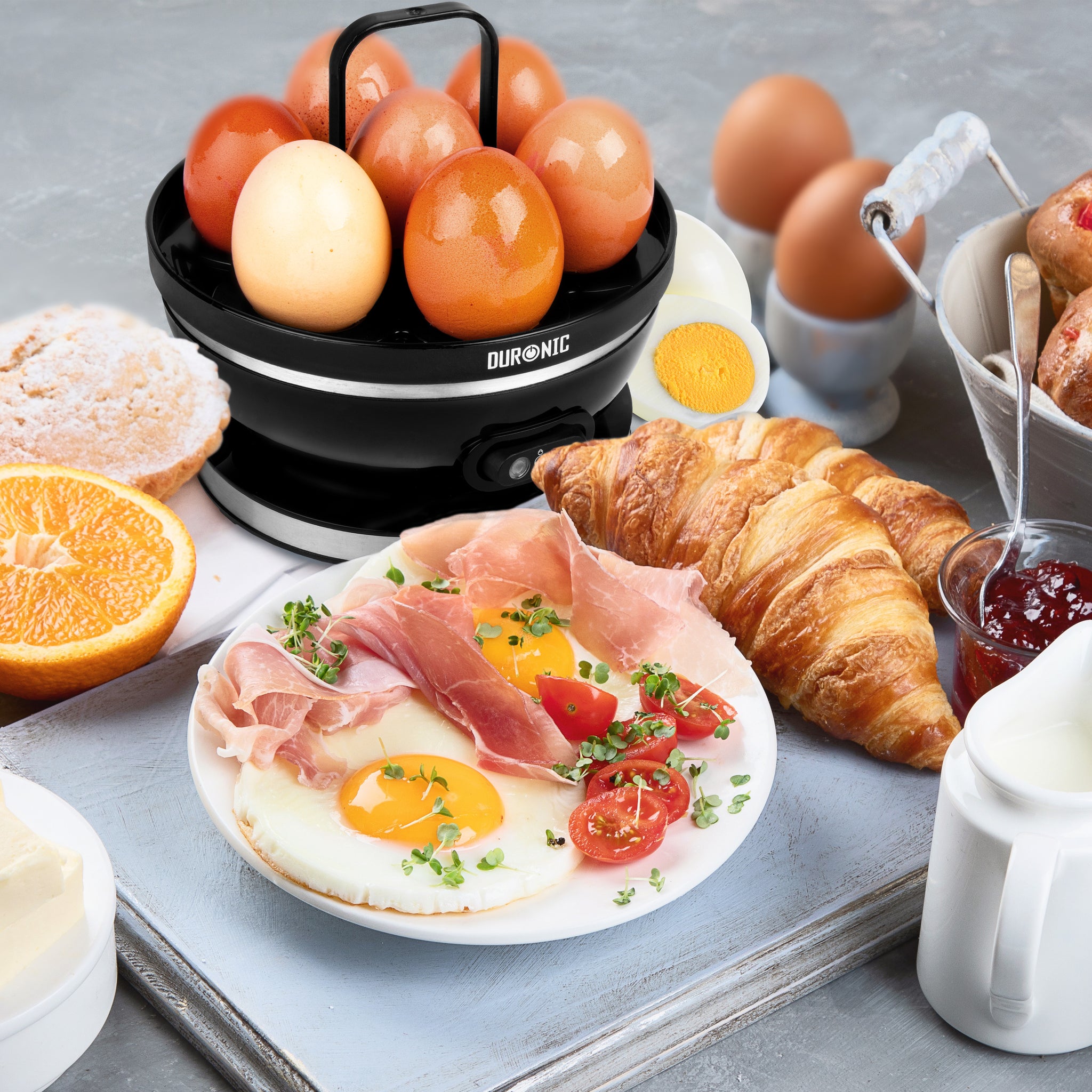 Duronic EB27 BK Hervidor de Huevos Eléctrico 400 W para 7 Huevos | Mostrando el hervidor de huevos eléctrico negro sobre una mesa con un desayuno completo que incluye huevos cocidos en su bandeja soporte, un plato con huevos fritos y jamón, croissants, fruta y mermelada
