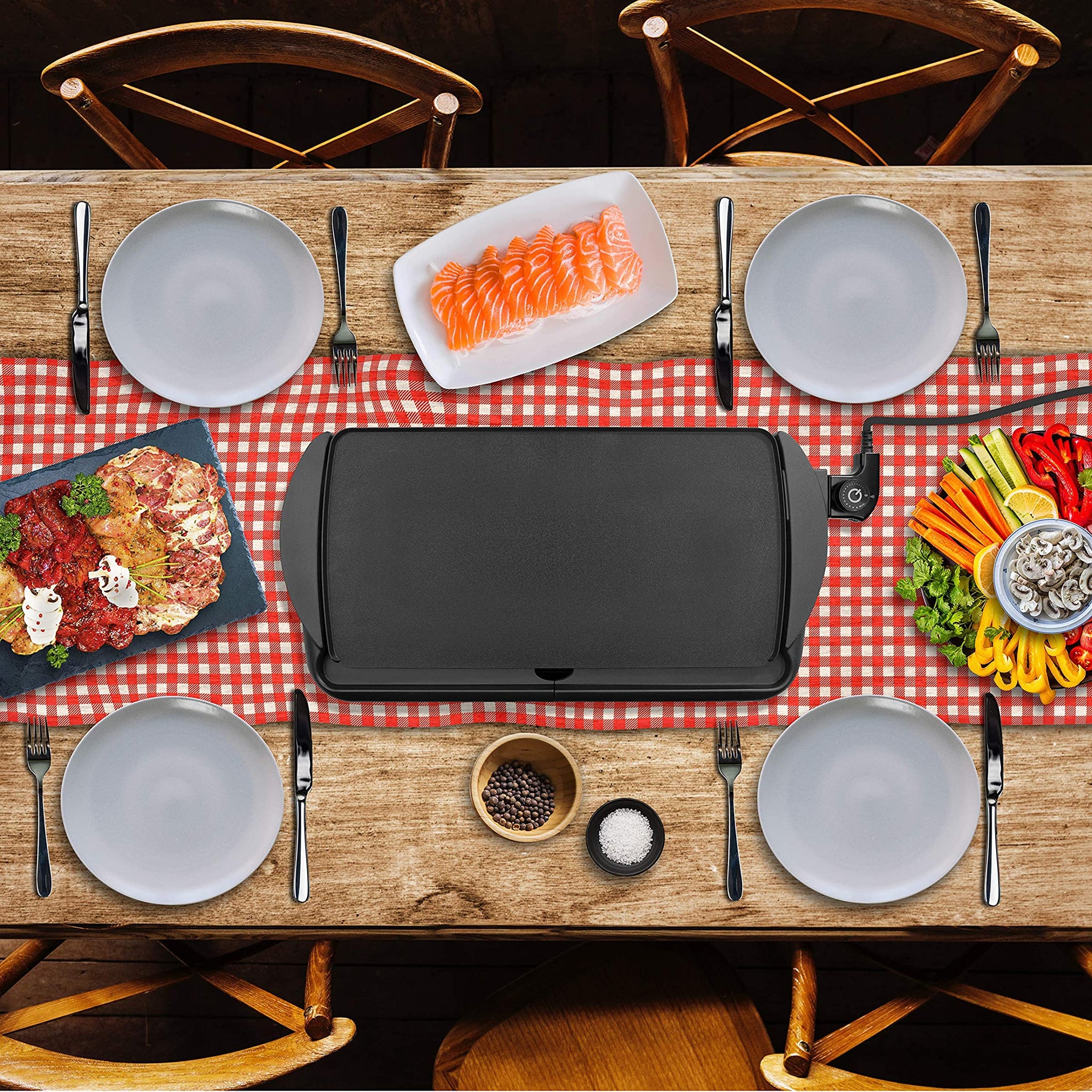 Duronic GP20 Plancha Eléctrica Grill Mostrando la plancha colocada en el centro de una mesa de comedor decorada con mantel de cuadros, rodeada de platos y diversos ingredientes frescos como salmón, carne y verduras listos para cocinar