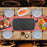 Duronic GP20 Plancha Eléctrica Grill Mostrando la plancha colocada en el centro de una mesa de comedor decorada con mantel de cuadros, rodeada de platos y diversos ingredientes frescos como salmón, carne y verduras listos para cocinar