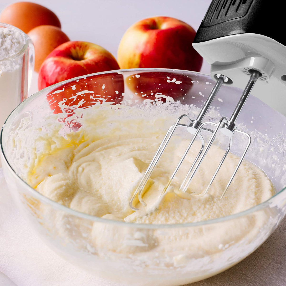 Duronic HM4 - Batidora de Varillas y Amasadora | Mostrando un primer plano de la batidora en funcionamiento, batiendo una mezcla clara dentro de un bol de cristal, con ingredientes de repostería como manzanas y huevos al fondo
